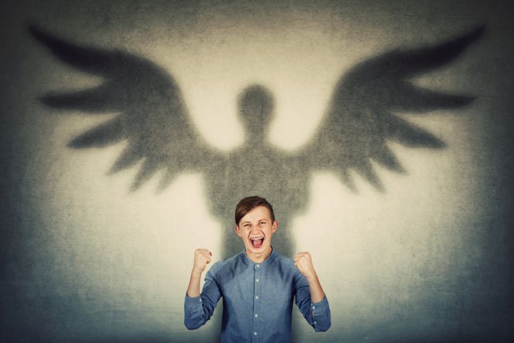 Furious teenage boy holding fists up and screaming showing his powers. Confident adolescent guy casting a superhero shadow with angel wings on a dark room wall. Inner strength and ambition concept.