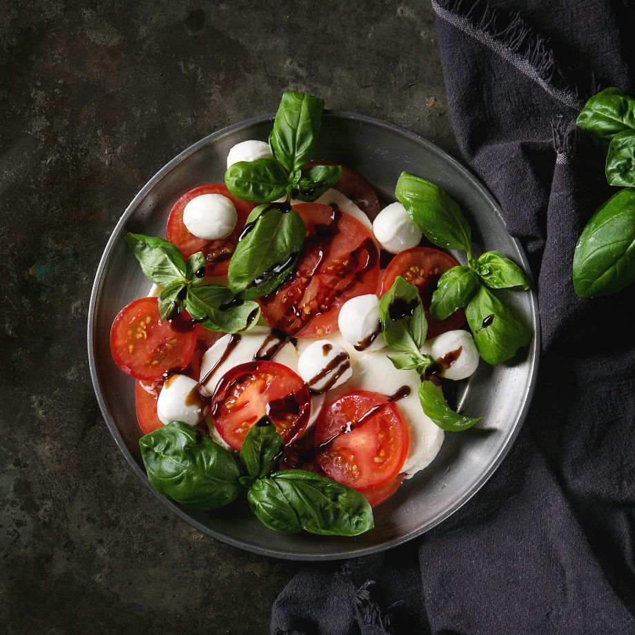 Italian caprese salad with sliced tomatoes, mozzarella cheese, basil, olive oil. Served in vintage metal plate on textile napkin over dark metal background. Top view. Rustic style. Square image
