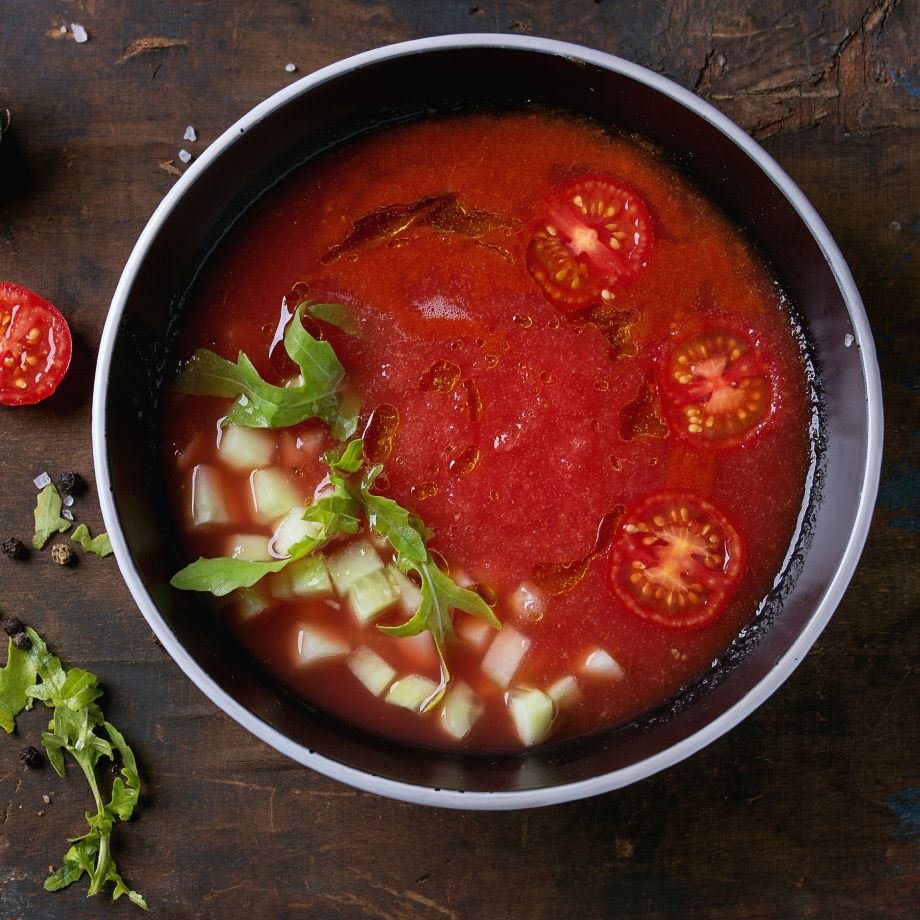 Black bowl of watermelon and tomato gazpacho cold soup, served with chopped cucumber, arugula and sliced cherry tomatoes over old wood background. Top view. Summer eating concept. Square image