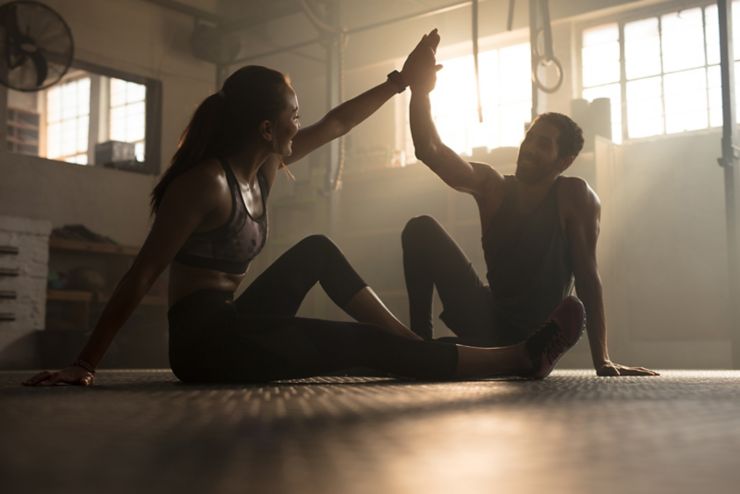 Healthy man and woman sitting on floor and giving each other high five at the gym. Fitness people after successful exercising session in gym.