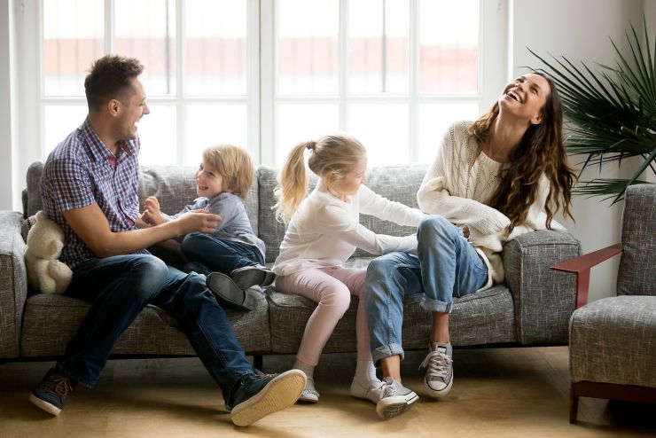 Happy parents and kids having fun tickling sitting together on sofa, cheerful couple laughing playing game with little active children son and daughter in living room at home, family funny activity