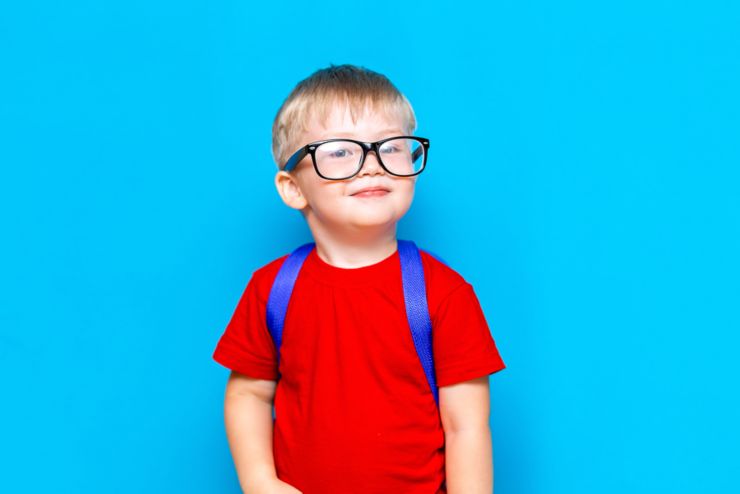 Happy smiling boy in red t-shirt with glasses on his head is going to school for the first time. Child with school bag. Kid on blue background background. Back to school.