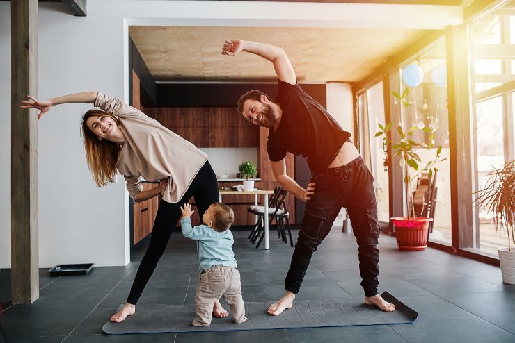 The family practices yoga, family life in isolation. Mom and dad are doing exercises, and infant child creeps next to his parents. Sunset light from the windows. Family quarantine; Shutterstock ID 1690523587