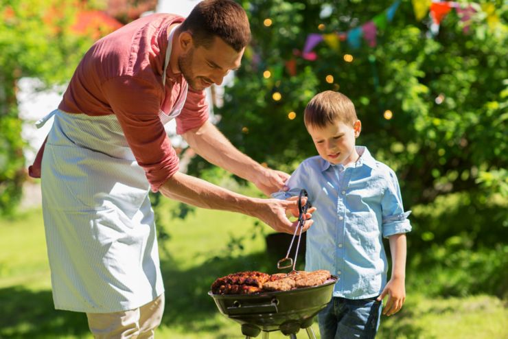 food, people and family time concept - father and son cooking meat on barbecue grill at bbq party in summer garden