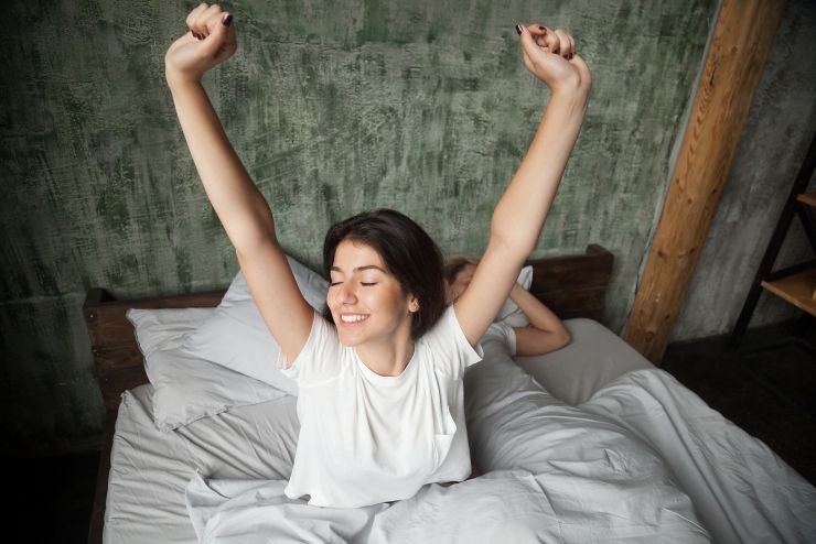 Young smiling woman waking up happy after healthy sleep stretching on comfortable bed with sleeping man at background, rested refreshed girl enjoys pleasant wakeup, good morning in cozy loft bedroom