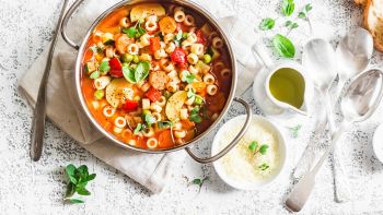 Minestrone soup in a pan on a light table, top view. Italian soup with pasta and seasonal vegetables. Delicious vegetarian food concept. Flat lay   